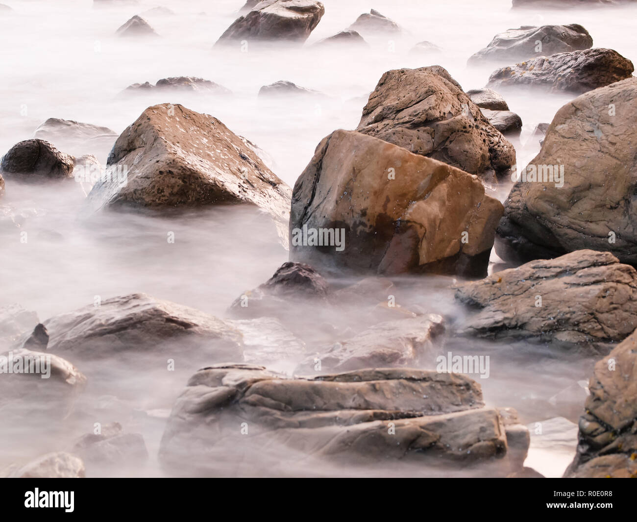 Slow exposure blurs the waves in the ocean as they break over rocks at sunset Stock Photo