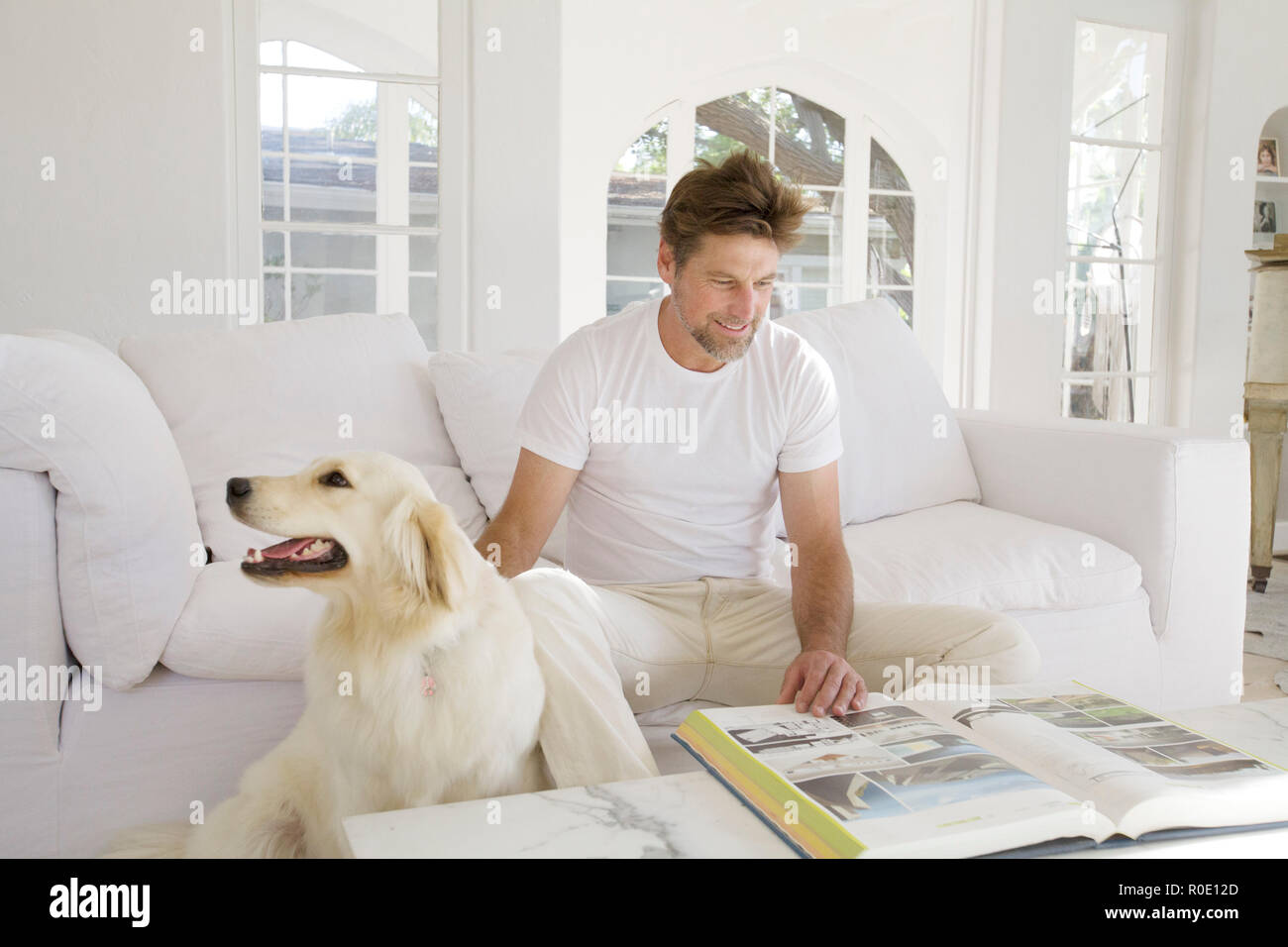 Mid-Adult Man Reading Large Book in Living Room while Petting Pet Dog I Stock Photo