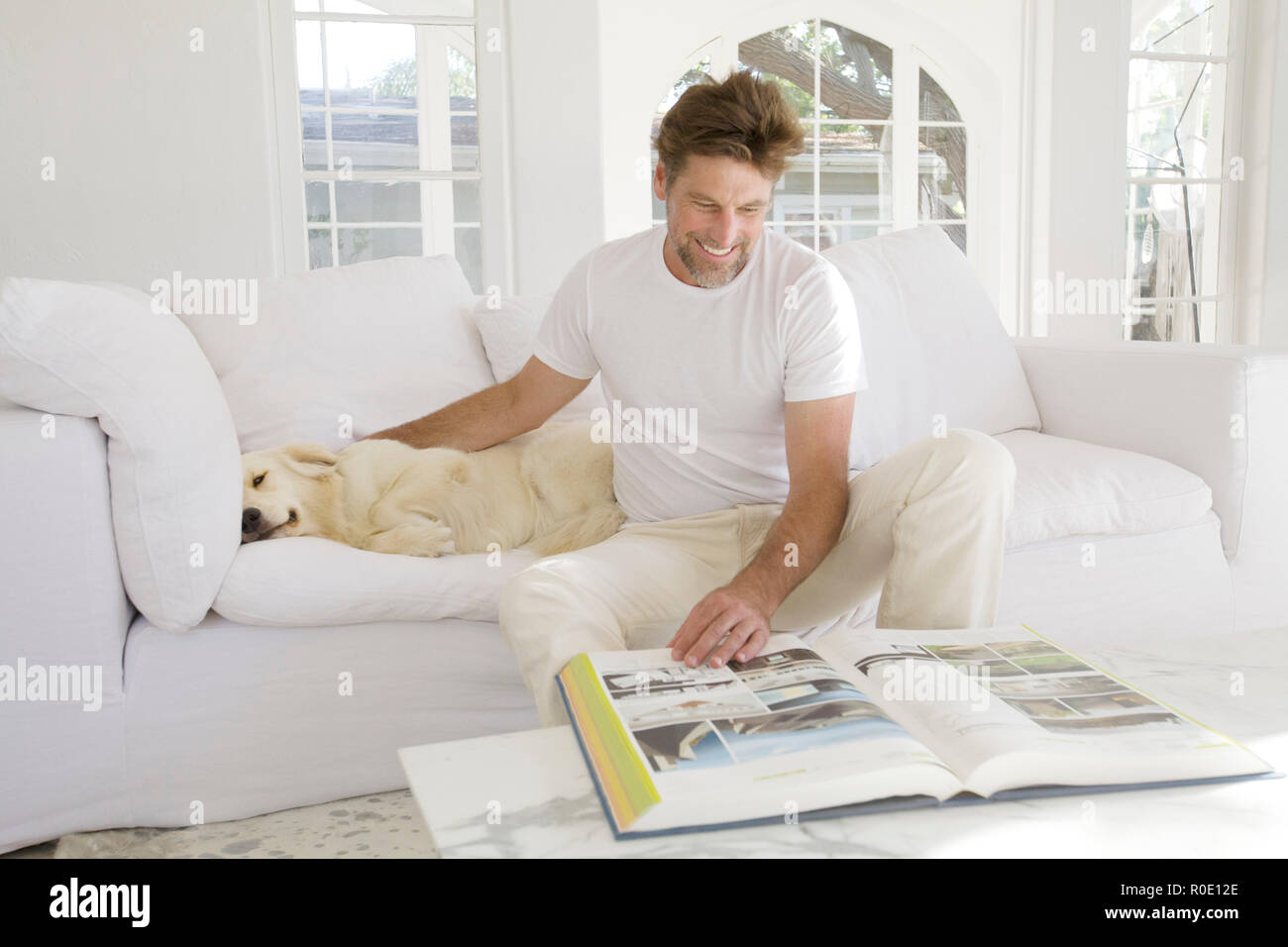 Mid-Adult Man Reading Large Book in Living Room while Petting Pet Dog II Stock Photo