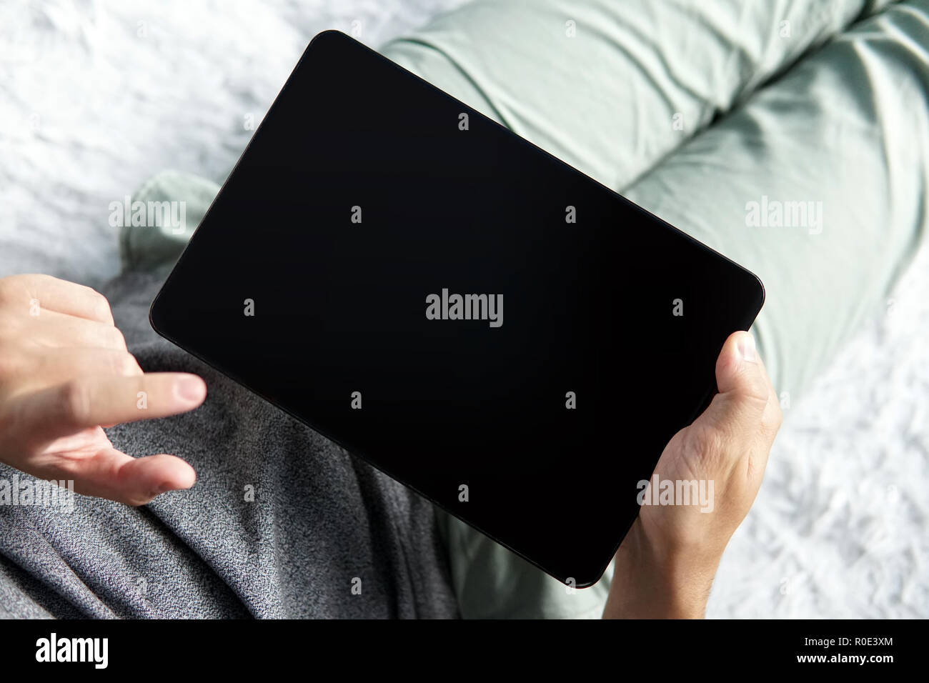 Close up of a young man comfortably lying on a bed with a tablet and touching the screen. Halfshot of a man sitting with black screen tablet in hands. Stock Photo