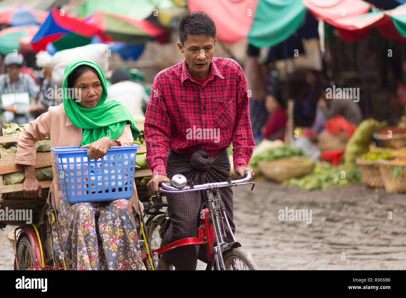 MANDALAY,MYANMAR,JANUARY 17, 2015 : A couple of vegetable seller is riding a sidecar bicycle in the muddy street of the Zegyo outdoors market, in Mand Stock Photo