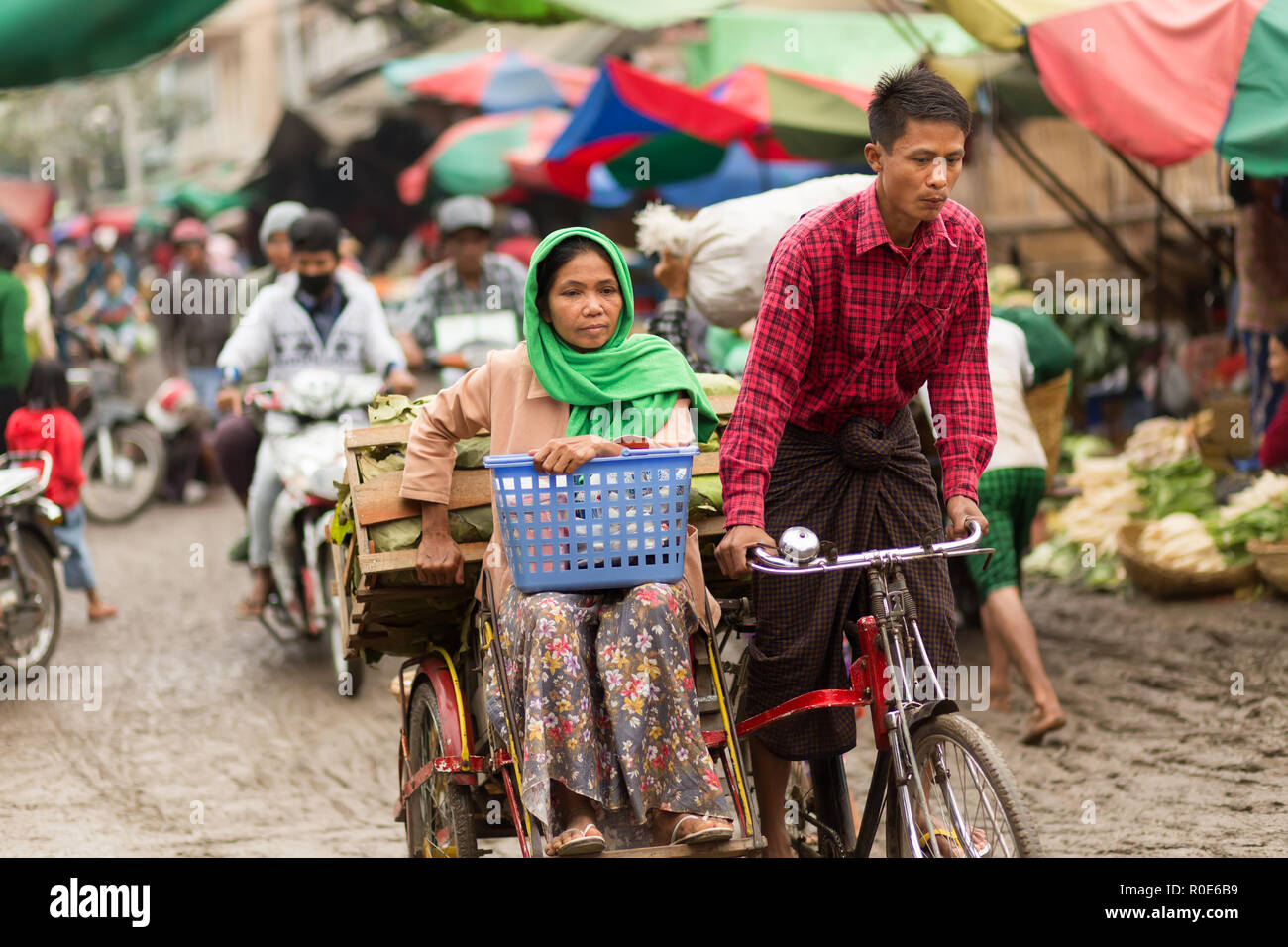 MANDALAY,MYANMAR,JANUARY 17, 2015 : A couple of vegetable seller is riding a sidecar bicycle in the muddy street of the Zegyo outdoors market, in Mand Stock Photo