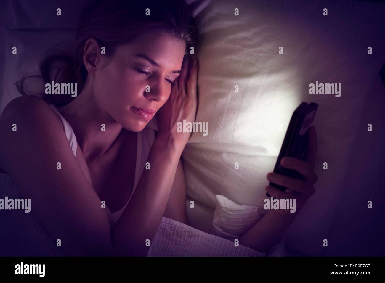 Young woman holding smartphone in bed and falling asleep Stock Photo