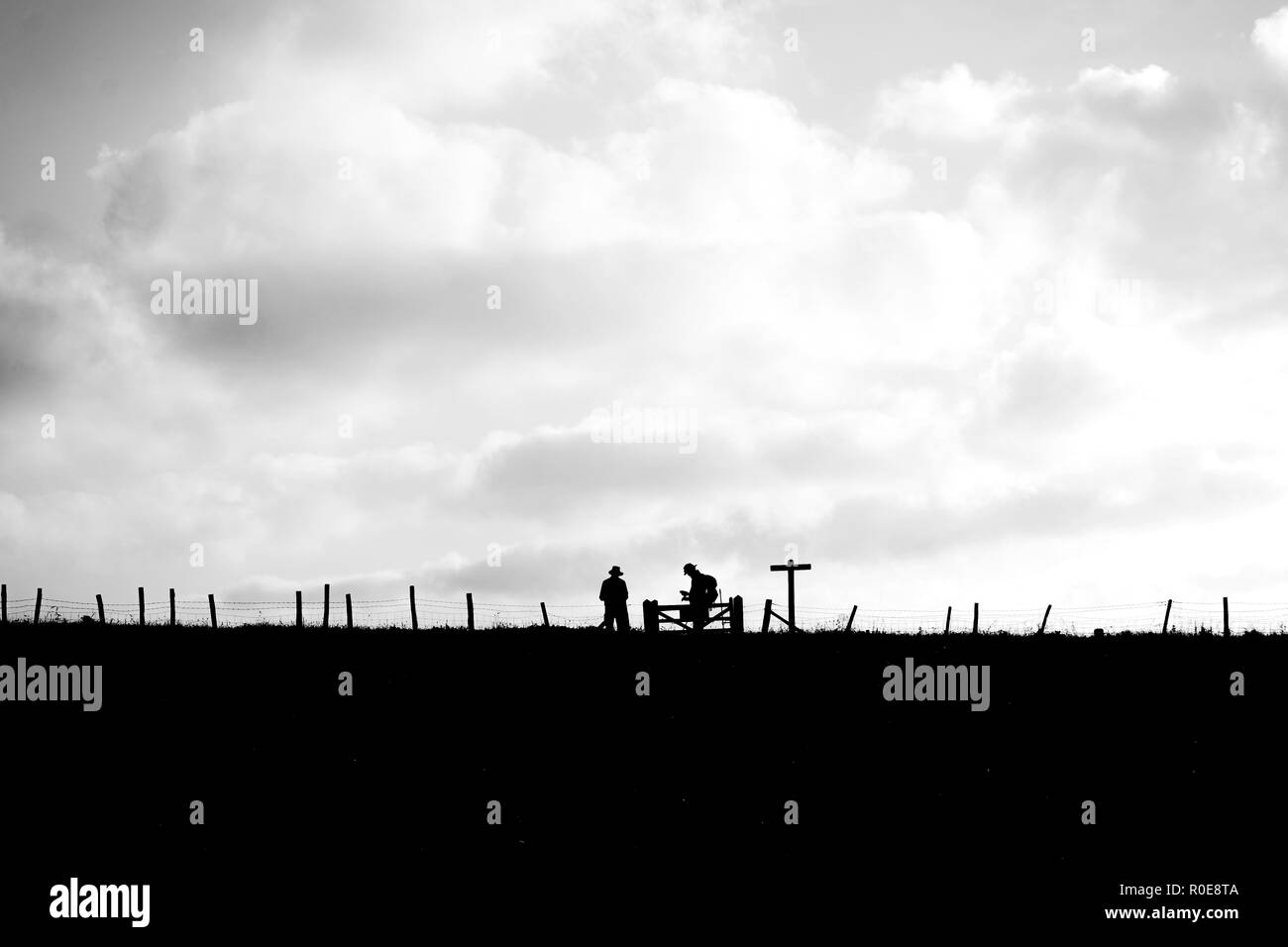 In the foreground is a field in the distance are two hikers silhouetted standing at a gate next to a sign post with a wooden fence running across the  Stock Photo
