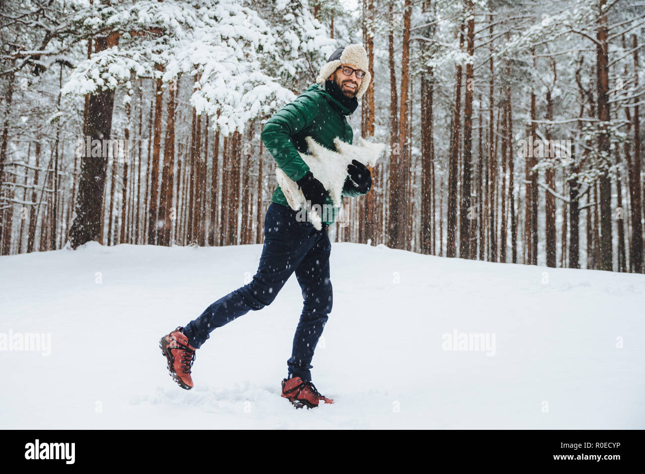 Outdoor portrait of positive happy male has fun, walks in winter forest, enjoys beautiful landscapes and fresh cold weather, holds artificial white fi Stock Photo