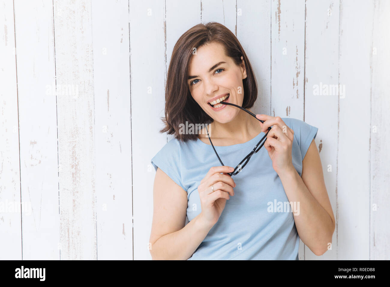Waist up portrait of blue eyed brunette female takes off spectacles, dressed casually, stands against white fence background, being in good mood. Chee Stock Photo