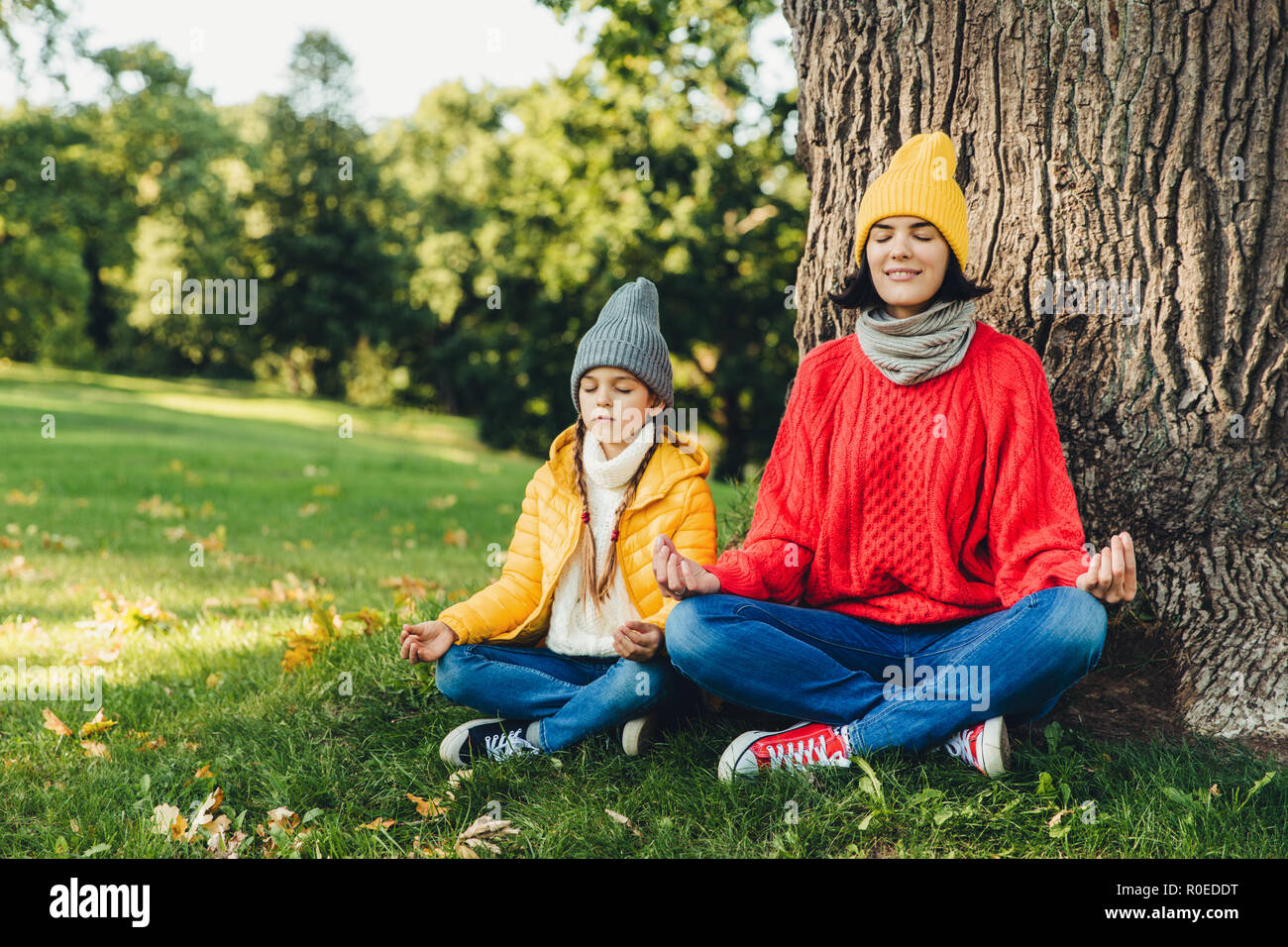 Relaxed carefree woman and little daughter sit in pose of lotus near tree in park, close eyes, try to concentrate, practice yoga together, dressed in  Stock Photo