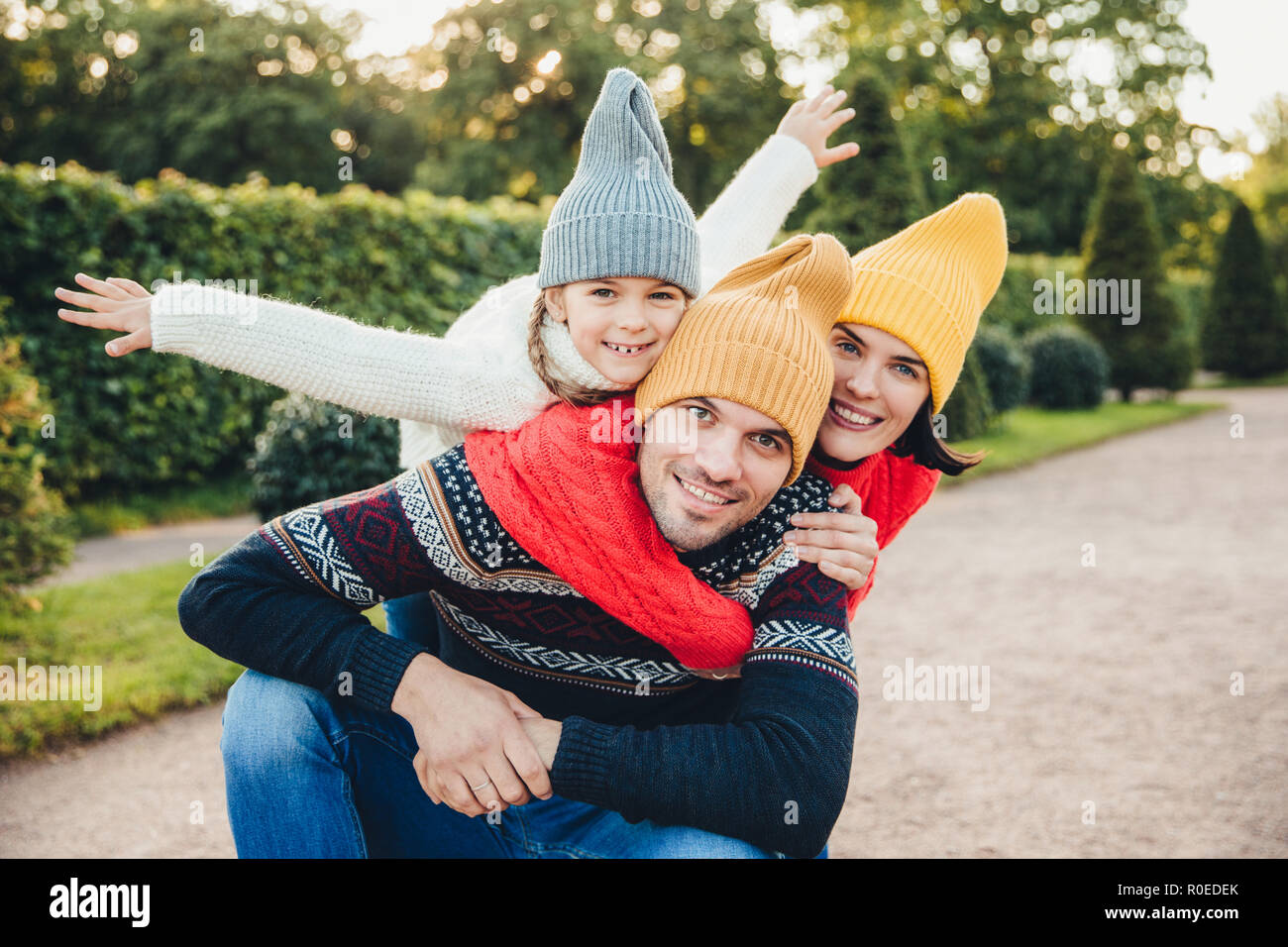 Having nice time together! Smiling excited woman, man and their little female child, wear warm knitted clothes, embrace each other, walk in park, bein Stock Photo