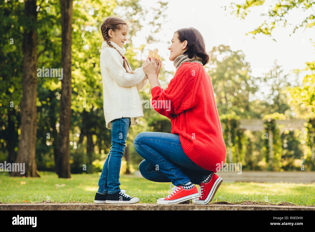 Young mother in warm knitted red sweater plays with her small daughter in park, gives her leaf, enjoy sunny autumn weather. Affectionate mom and littl Stock Photo