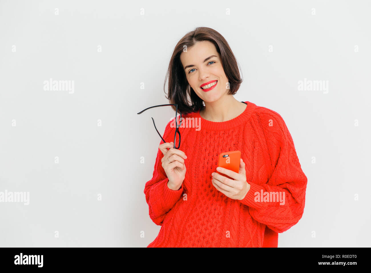 Pretty female with dark short hair, wears oversized red winter sweater, holds eyewear and smart phone, happy to message with friends, stands against w Stock Photo