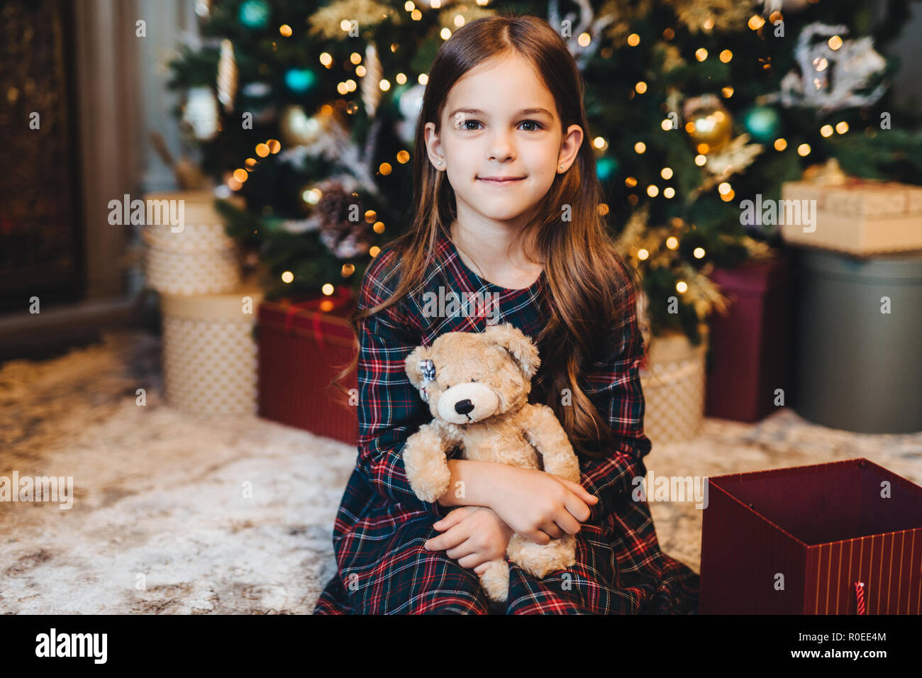 Small female child with appealing appearance, holds teddy bear, recieves present from parents, sits near Christmas tree. Adorable girl being glad to g Stock Photo