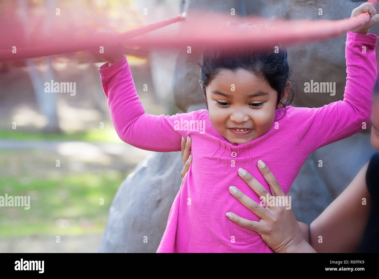 Pretty young girl of mixed race holding on to ropes, and excited as she walks on a balance beam at a kids playground while her mom helps to support he Stock Photo