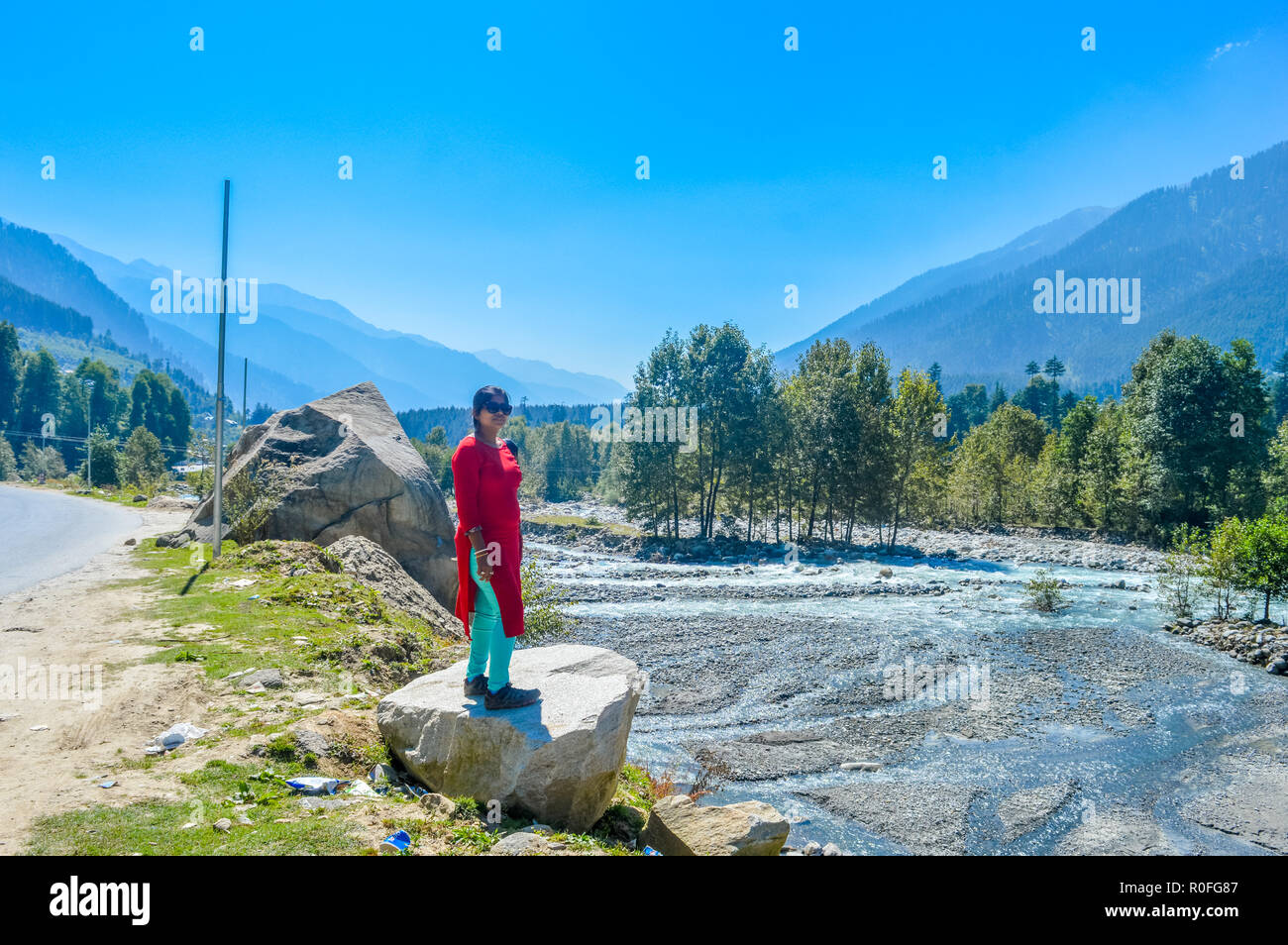 A lady in red drees standing on a bolder rock near a highway road. Beautiful landscape panorama view Himalayan valley of 'MANALI-LEH-ROAD, KULLU, JAMM Stock Photo
