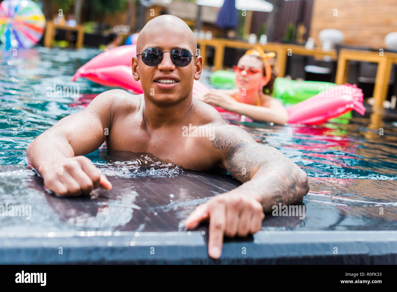young tattooed man in sunglasses looking at camera while his girlfriend  resting on inflatable mattress in swimming pool Stock Photo - Alamy