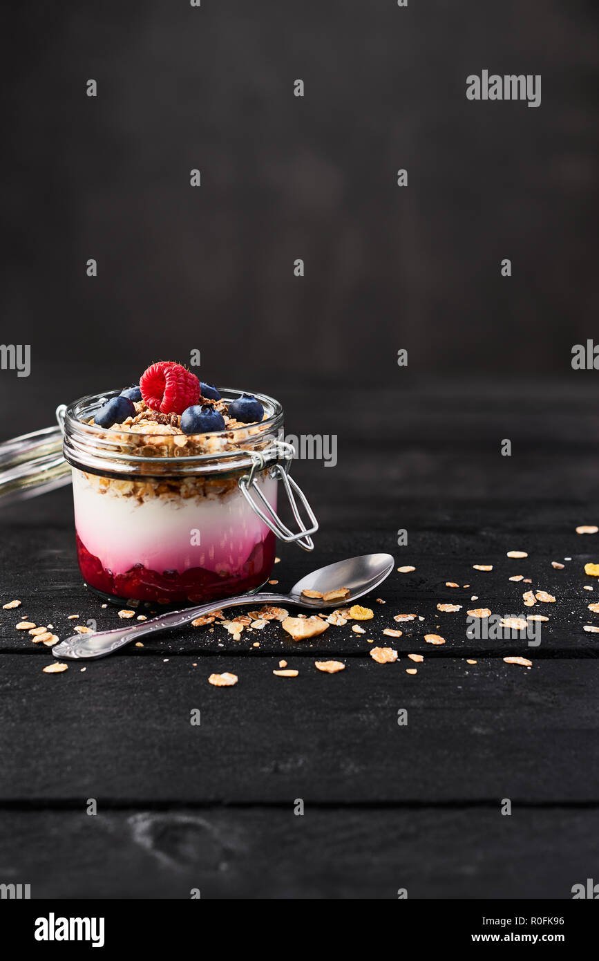 Healthy breakfast. Jar of healthy yogurt with raspberry sauce, oat and blueberries on black wooden table over dark concrete background with copy space Stock Photo