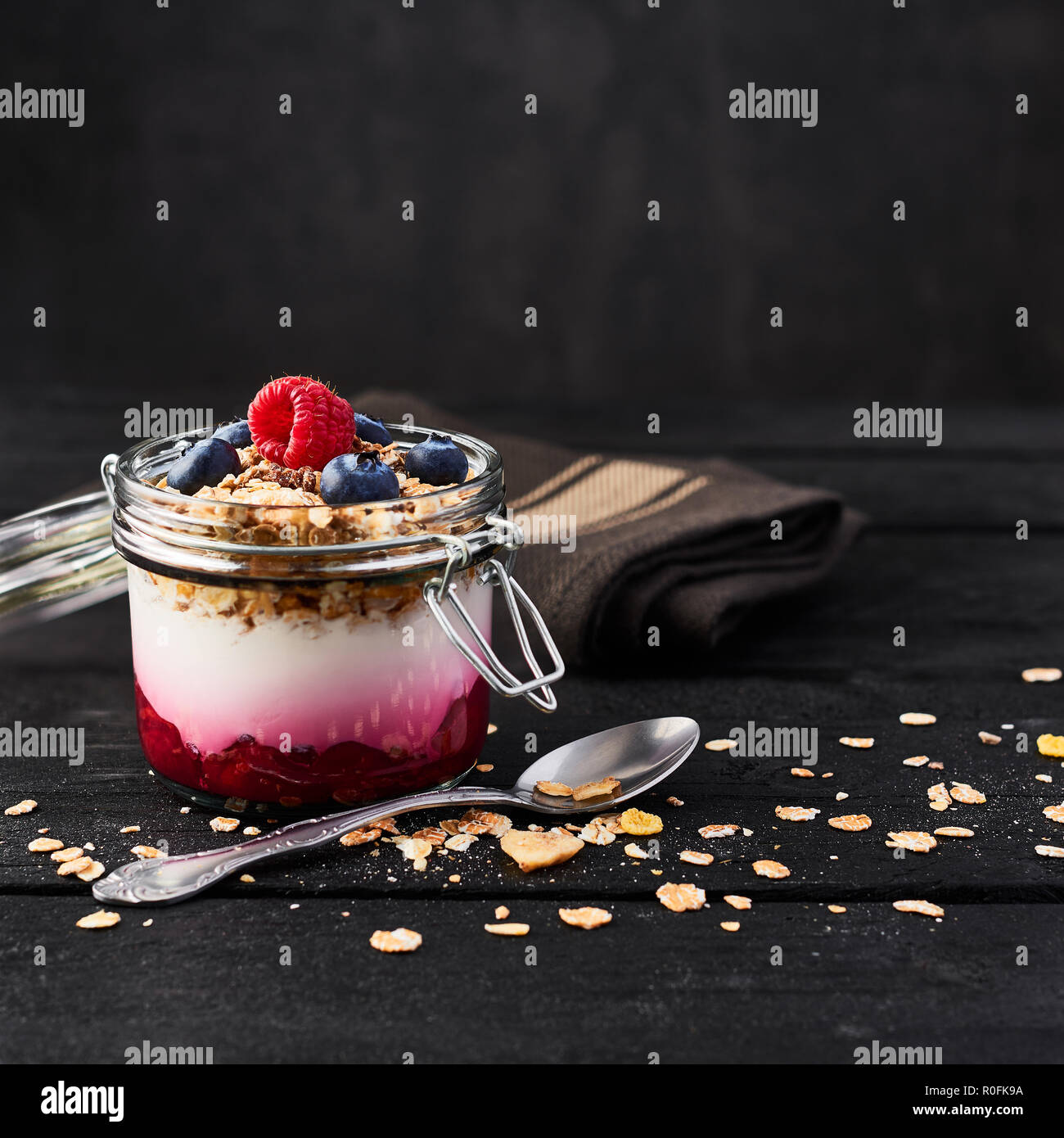 Healthy breakfast. Jar of healthy yogurt with raspberry sauce, oat and blueberries on black wooden table over dark concrete background with copy space Stock Photo