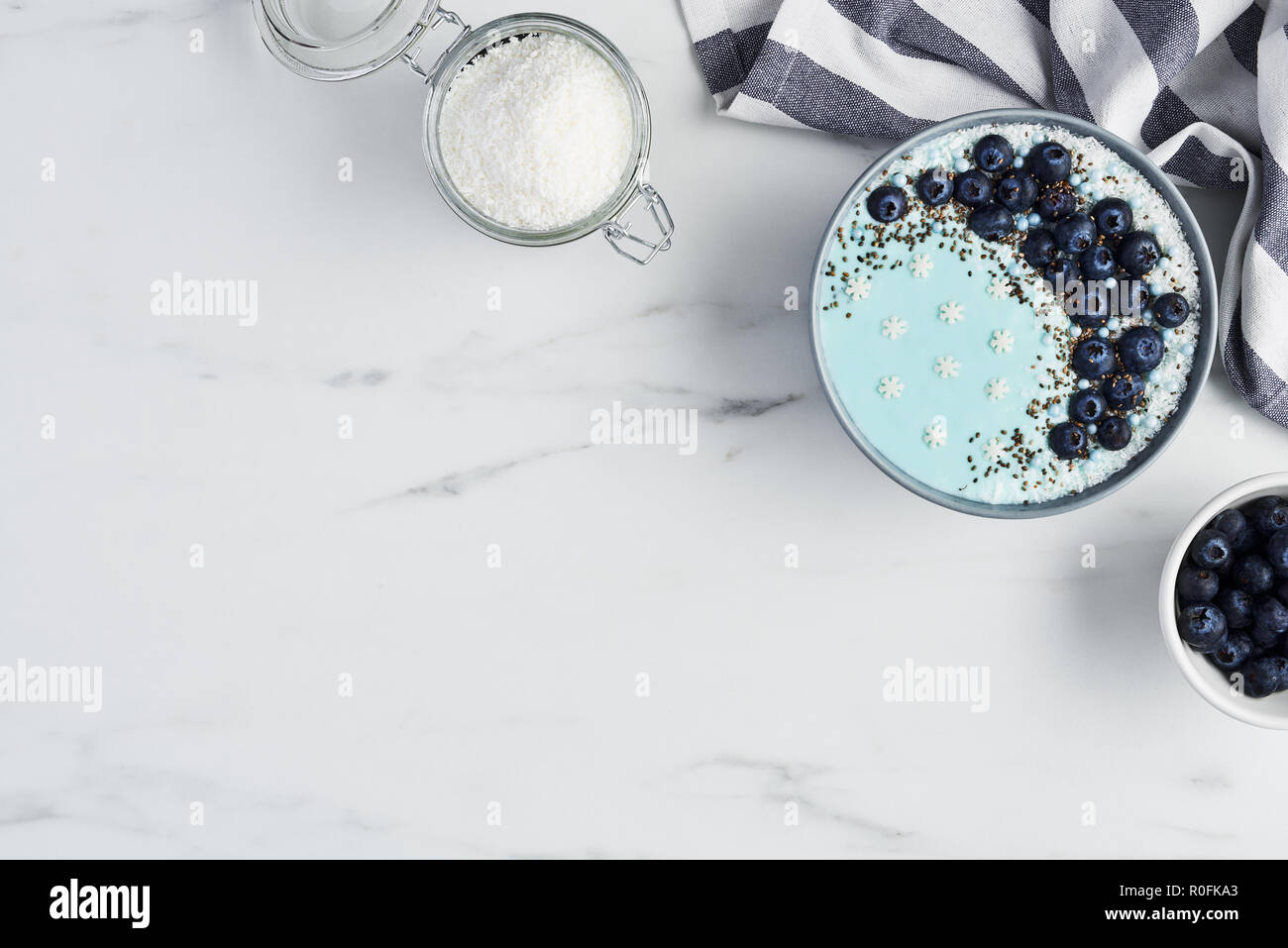 Top view of blue yogurt smoothie bowl made with blueberry, coconut flour, chia seeds and sugar pearls on white marble table with copy space. Stock Photo