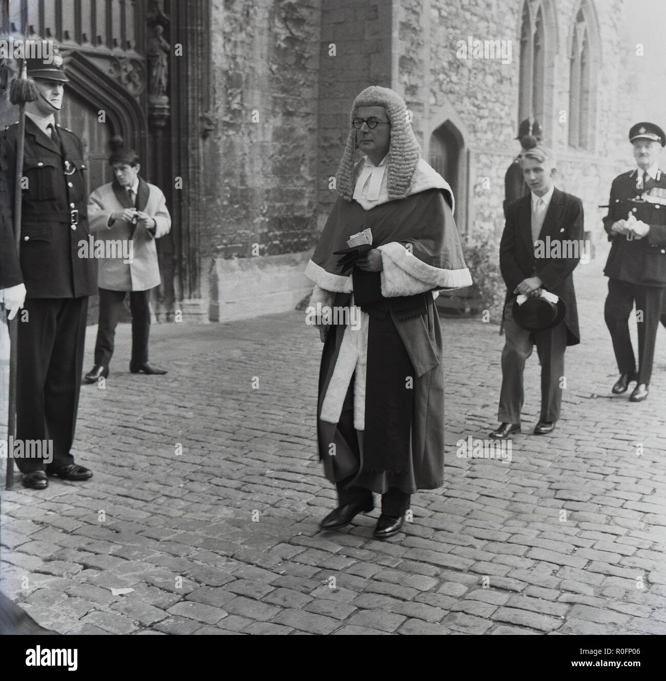 1964, Battle of Britain parade, local civic dignitaries, including those from the church of England, the Chief Constable and the judiciary - seen here in his long robes and wig -  walk in an orderly fashion along a cobbled path outside a church with a policeman in attendance, England, UK. Such parades or processions were took place in  English market towns in this era to remember the sacrficies the British military and public made during WW2. Stock Photo