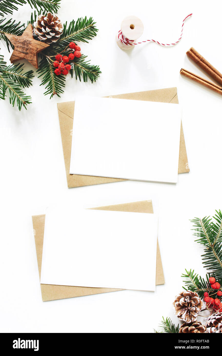Christmas composition. Blank greeting card, envelope mock-up scene. Pine cones, fir tree branches, red rowan berries and wooden stars on white table background. Flat lay, top view, vertical. Stock Photo