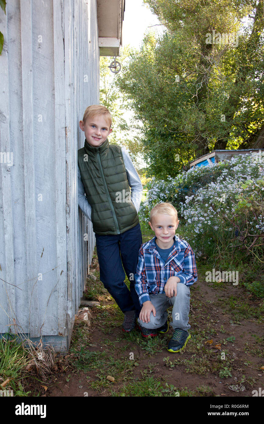 Two cute blond children lean casually against a shed or barn in autumn ...