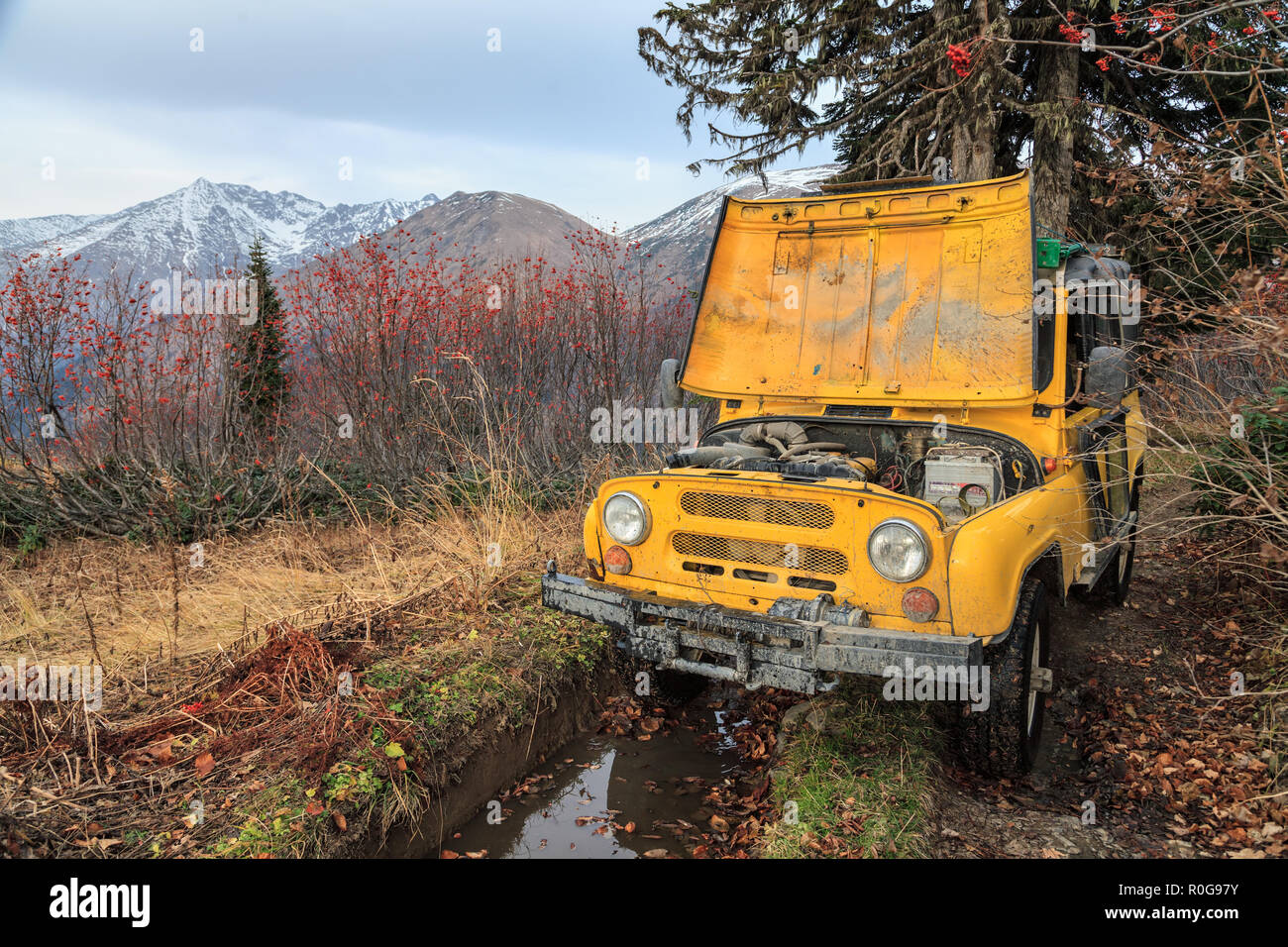 Yellow offroad all terrain car after sudden breakdown under repair on mountain forest road and mountain peaks background. Extreme outdoor activities.  Stock Photo
