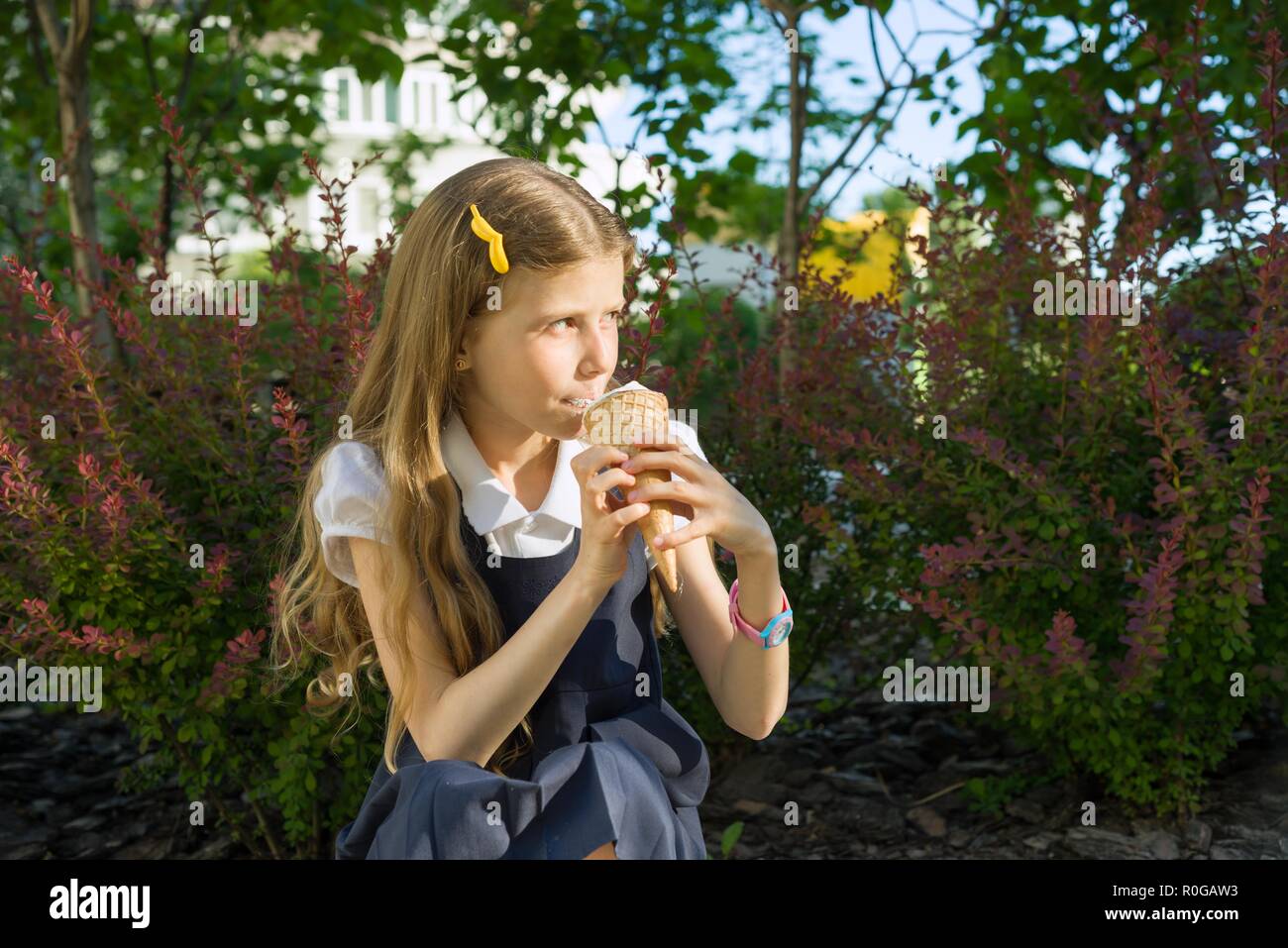 Schoolgirl 8 years old eating ice cream. Blond girl in school uniform, background of green trees, yard. Stock Photo
