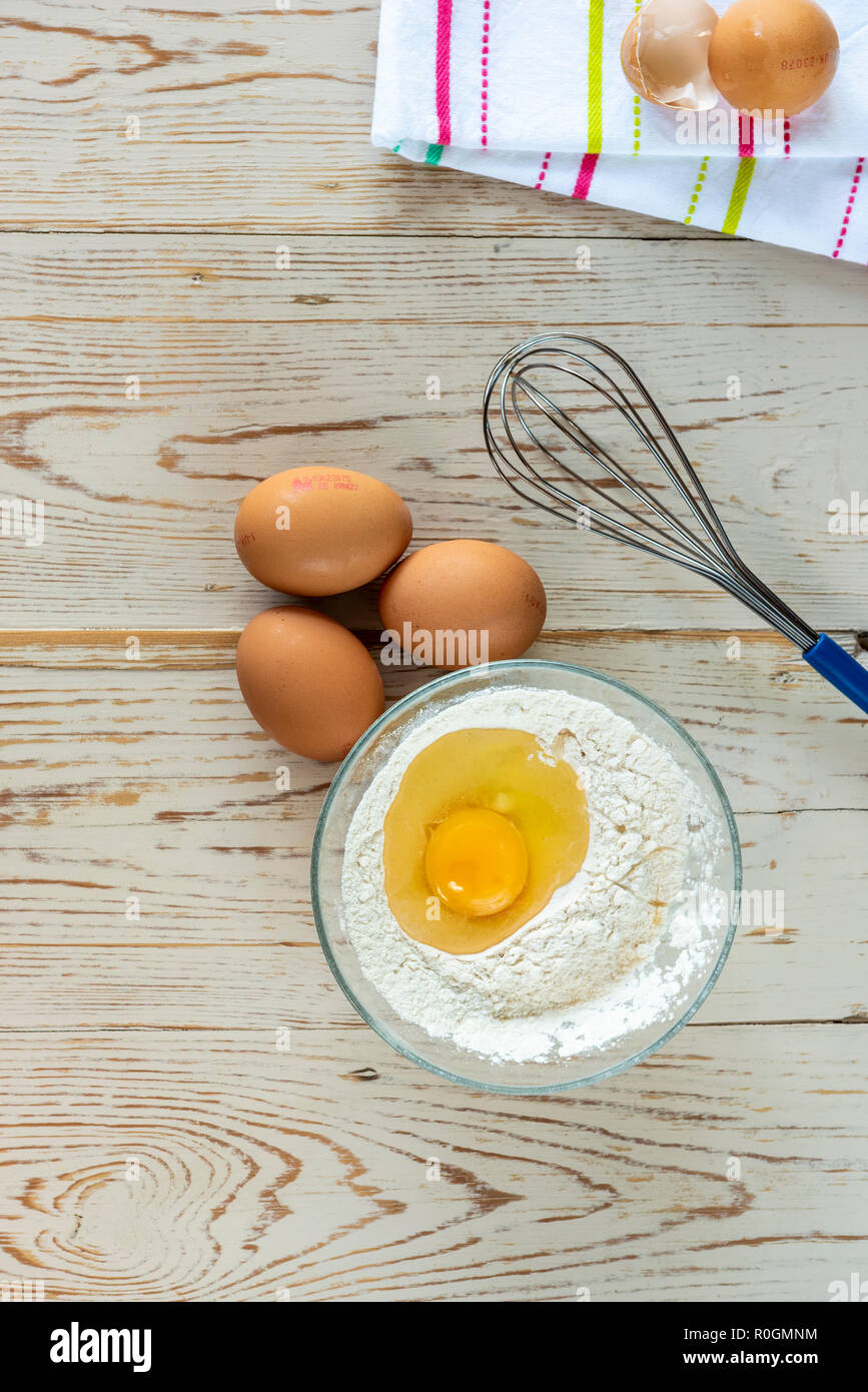 Baking ingredients, flour eggs with whisk, with broken eggshell on tea towel. Stock Photo