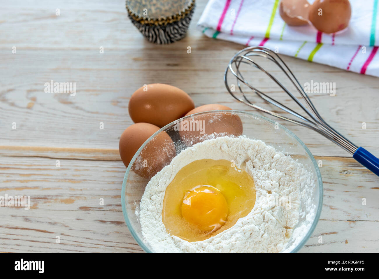 Baking ingredients, flour eggs with whisk, with broken eggshell on tea towel. Stock Photo