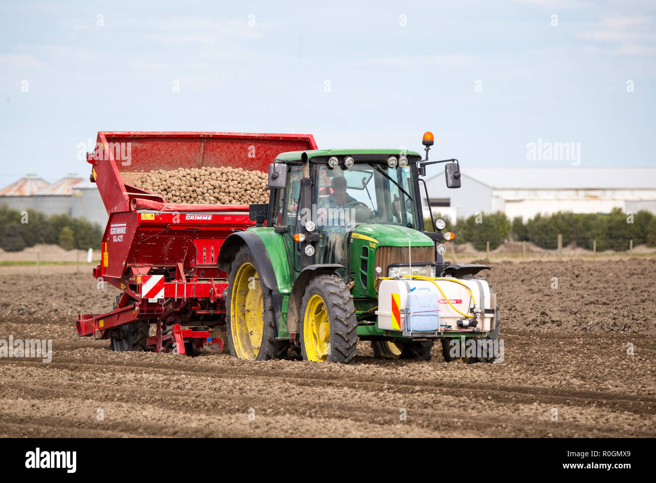 A farm worker uses a Gimme potato seeder and John Deere tractor in a farm field to plant seed potatoes in spring Stock Photo