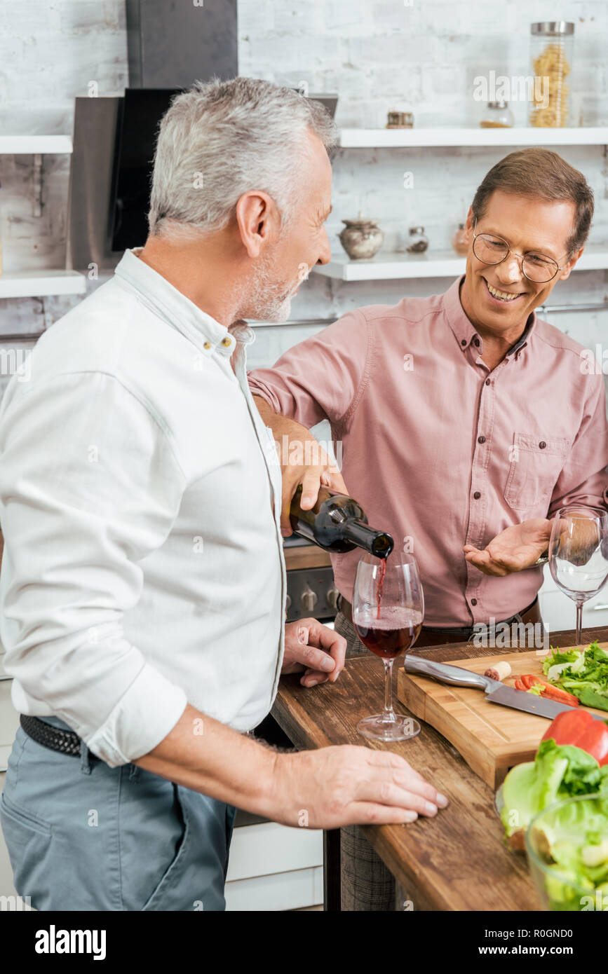 handsome happy man pouring wine to old friend during dinner in kitchen ...