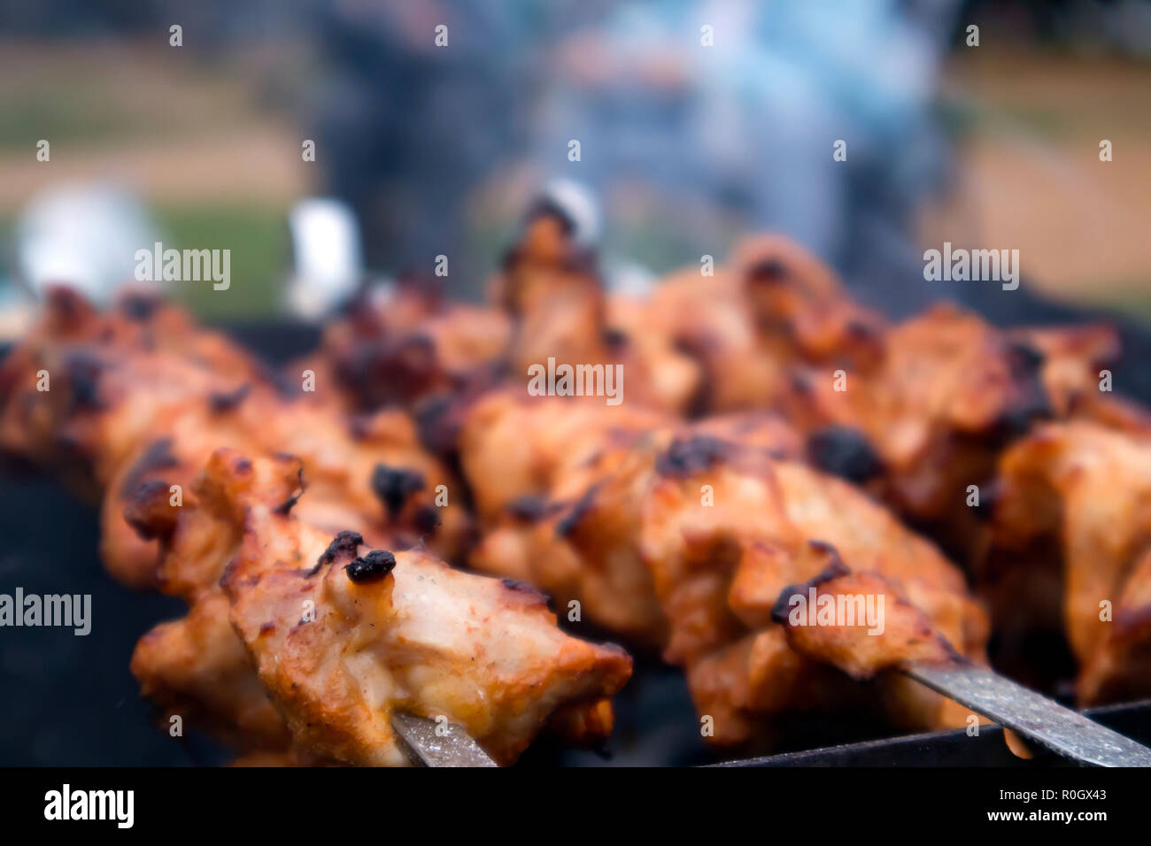 Skewers with roasted chiken meat on the hot grill closeup with two blurred human silhouettes in the background Stock Photo