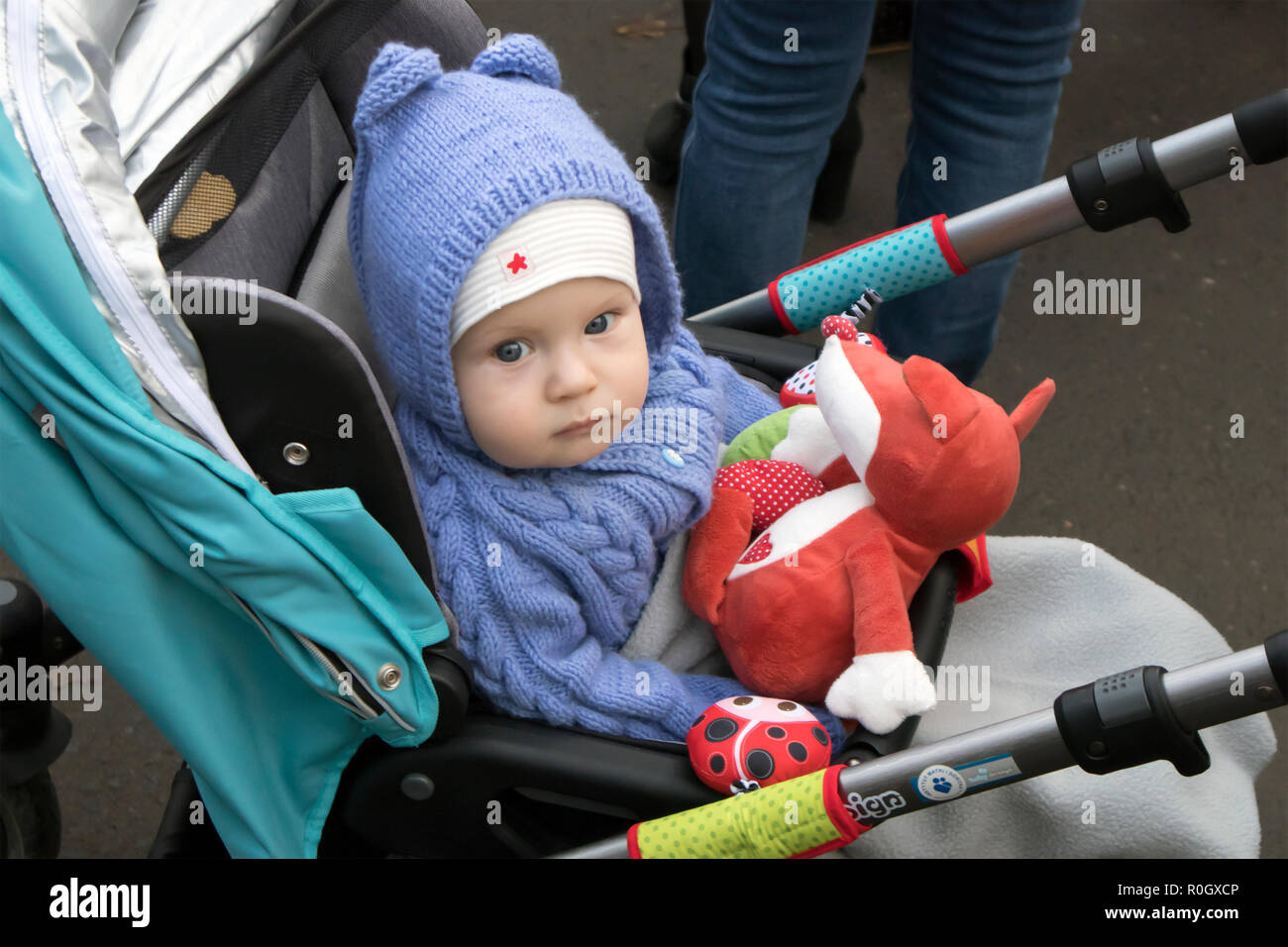 Cute serious baby sitting in stroller dressed in knitted azure costume with red toy in hands Stock Photo