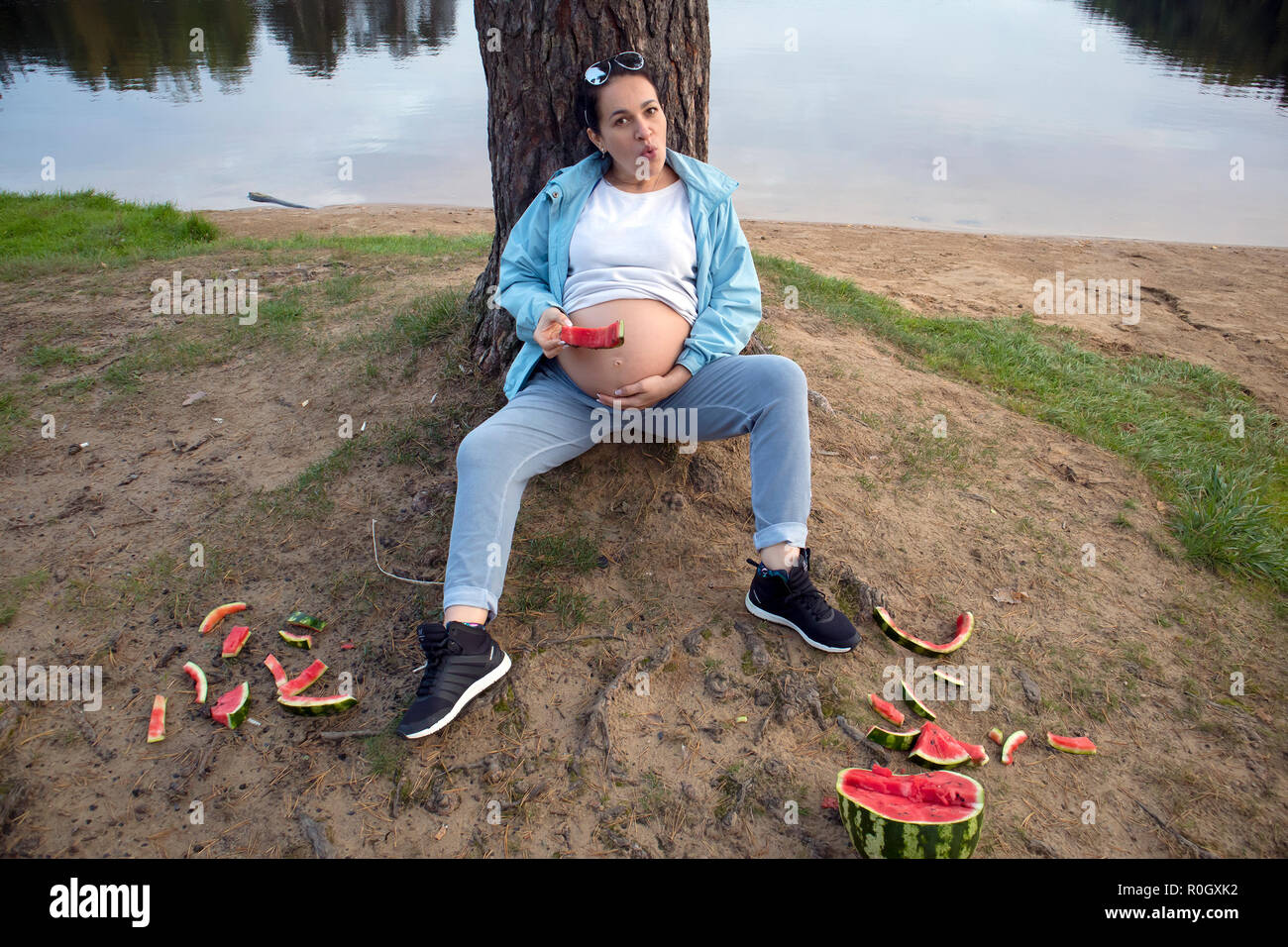 Attractive pregnant brunette with watermelon and scattered watermelon peels posing near the forest lake Stock Photo