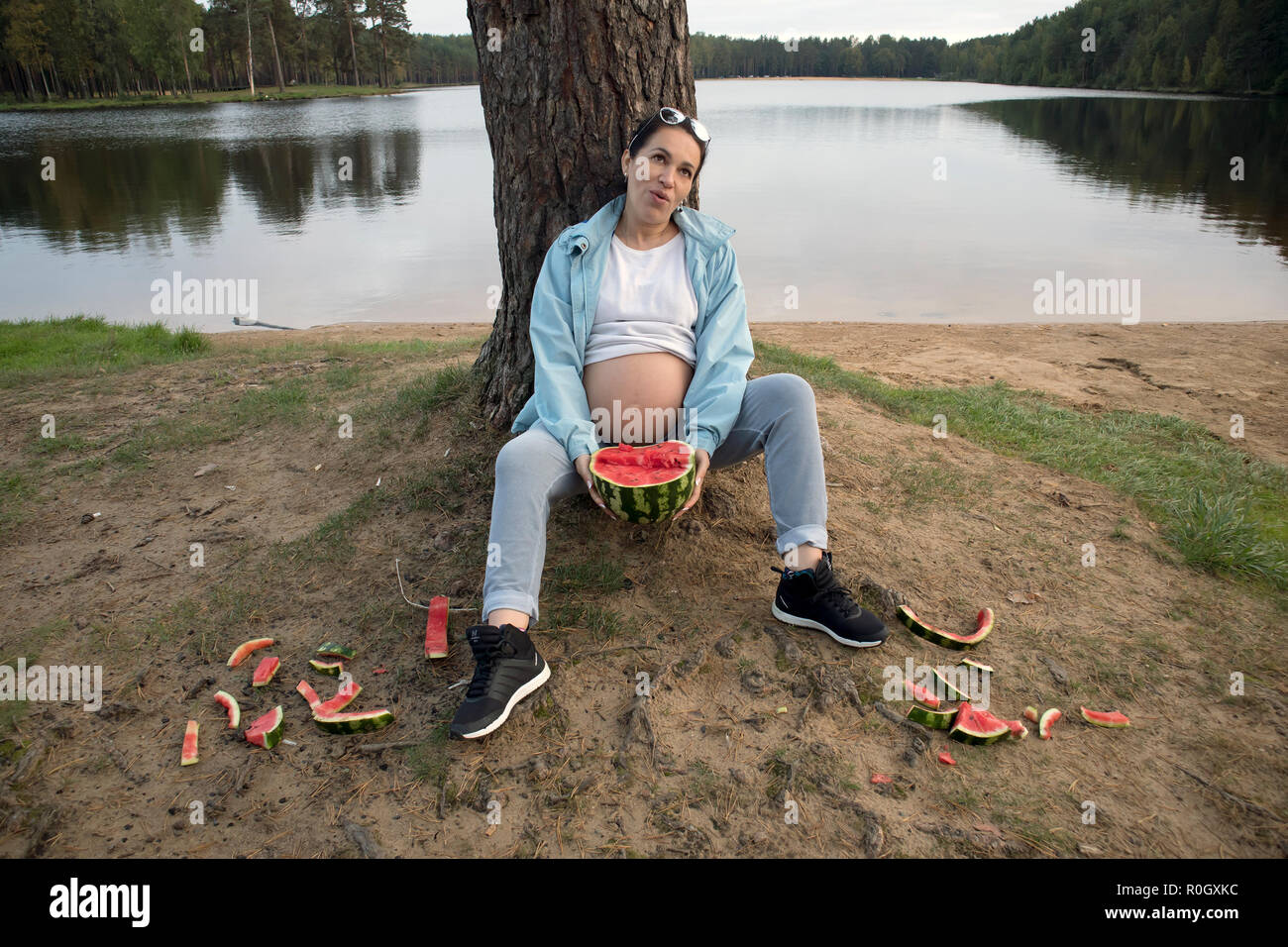 Attractive pregnant brunette with watermelon and scattered watermelon peels posing near the forest lake Stock Photo