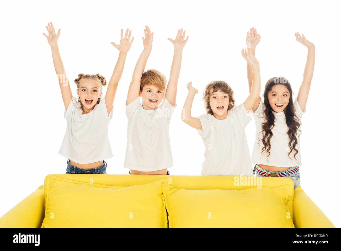 happy children standing behind yellow sofa with raised hands isolated on white Stock Photo