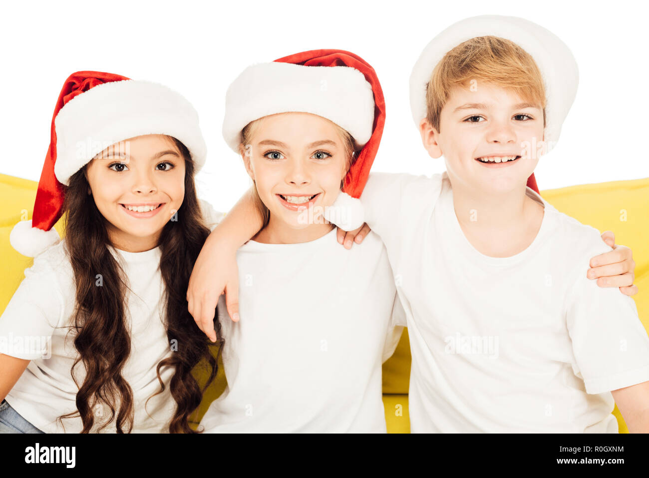 portrait of smiling adorable kids in santa hats sitting on yellow sofa and looking at camera isolated on white Stock Photo