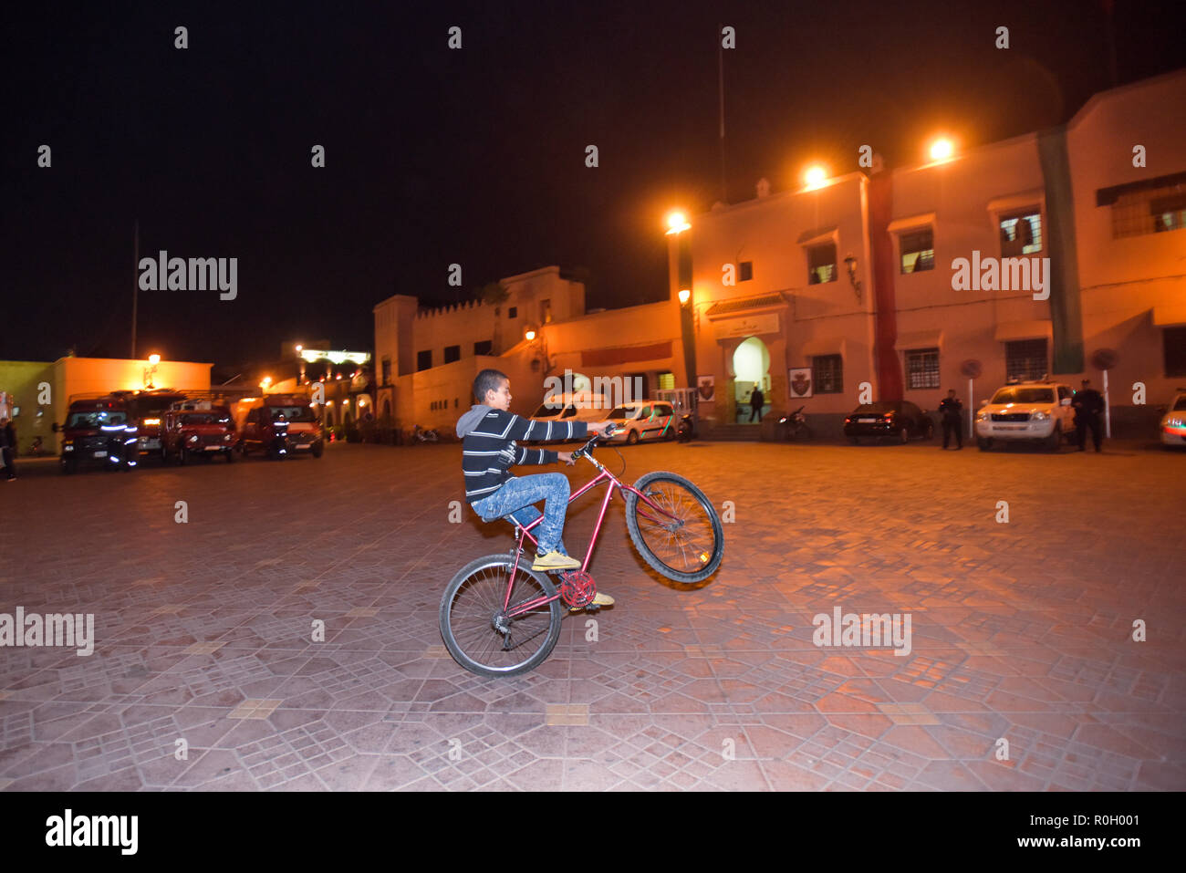 Lonely teenager boy at night playing with his mountain bike on one wheel in front of Marrakesh Police station Morocco North Africa Stock Photo