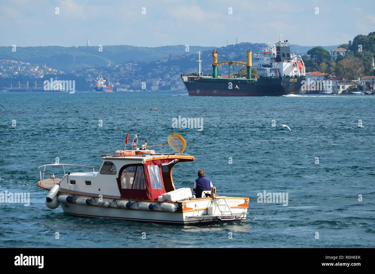 Large cargo ship goes along the Bosphorus, a small fishing boat with a fisherman in the foreground. Stock Photo