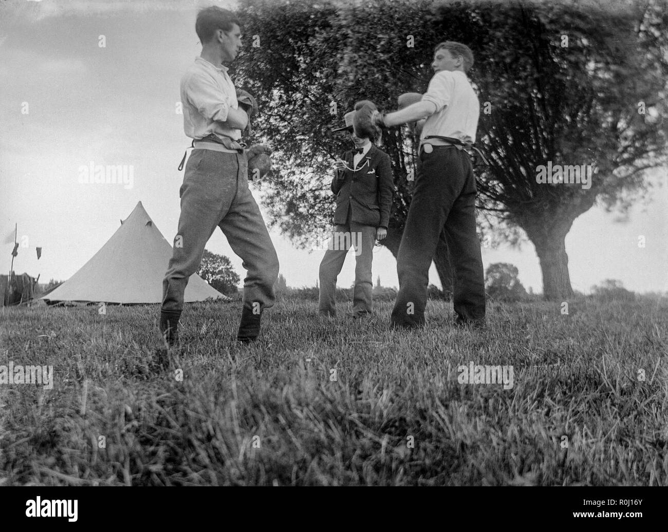 Edwardian black and white photograph showing two boys, or young men, on a camping trip in a field in England, squaring up for a boxing match, complete with vintage boxing gloves. A third boy acting as referee and timekeeper. Stock Photo