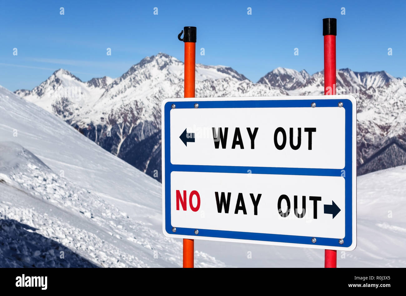 Way out vs no way out information direction sign bifurcating streams at ski resort against snowy mountain peak and blue sky winter background. Stock Photo