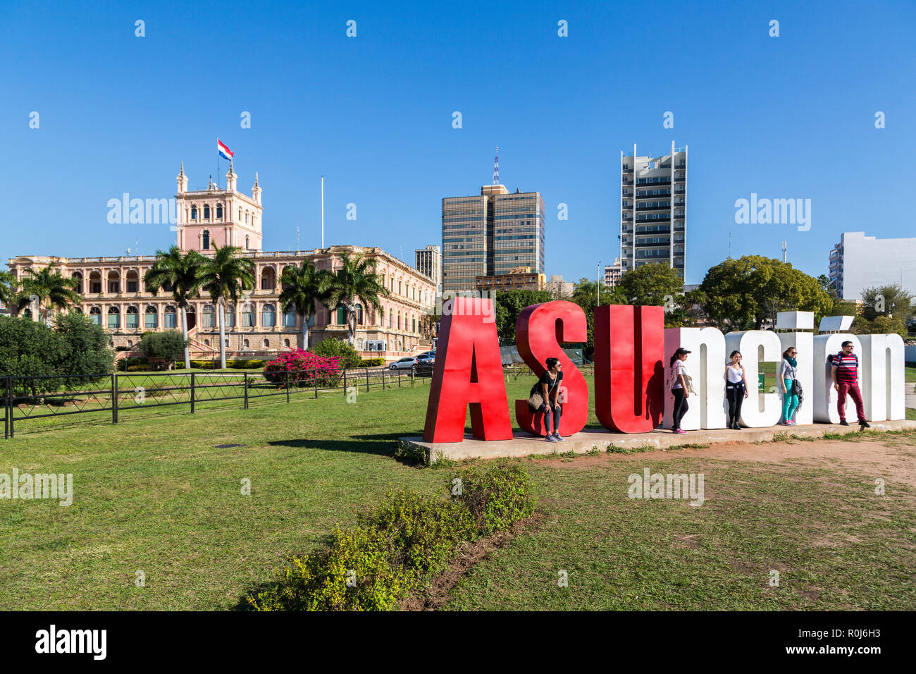 Five tourists are posing with 'ASUnción' letters and Palacio de los López (Presidential palace) in background. Asuncion city, Paraguay, Latin America Stock Photo