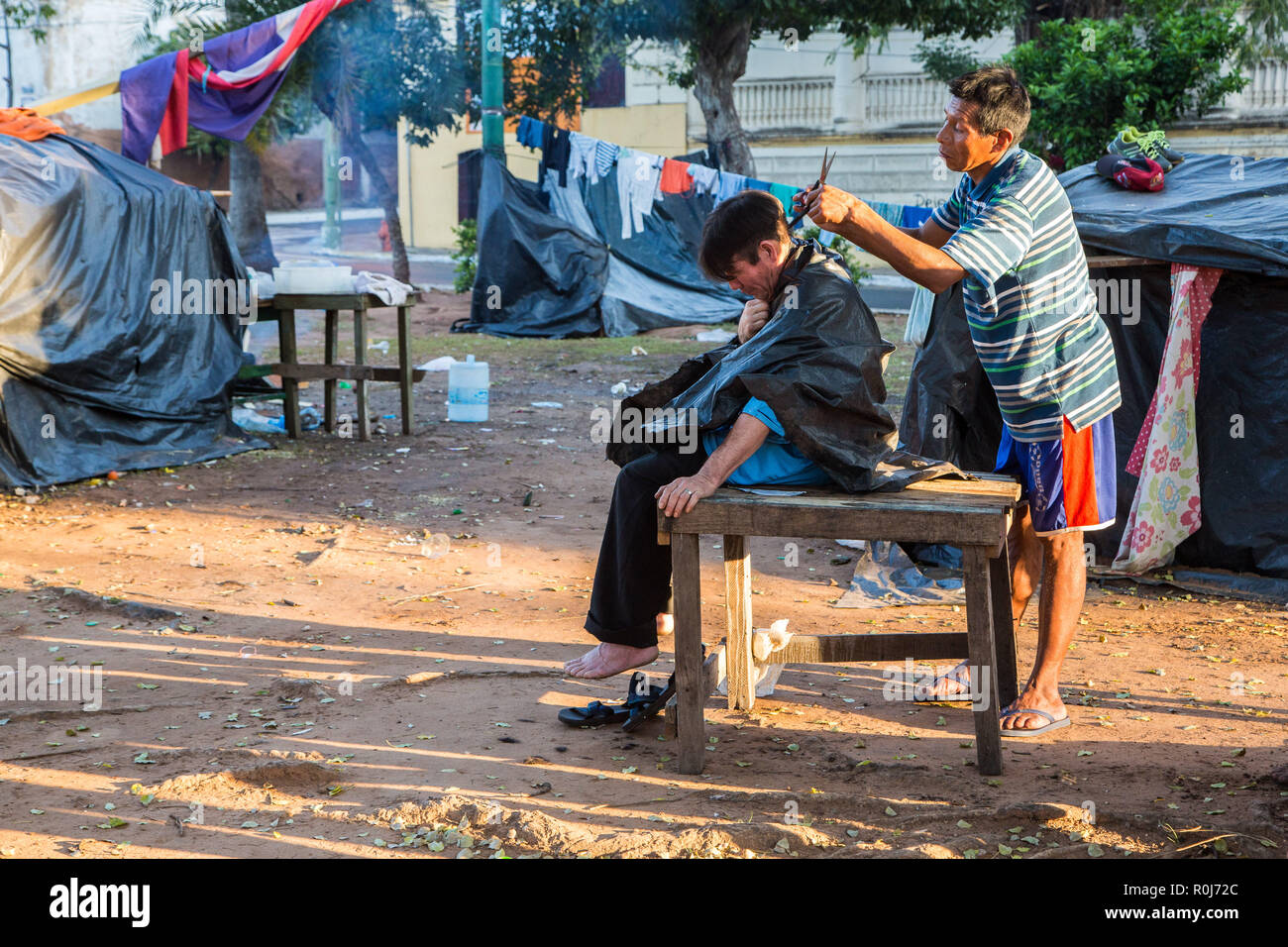 Outdoor haircut in slums of Asuncion city. Hairdressing on streets of Ciudad de Asunción Paraguay. Two men, tent camp. Stock Photo