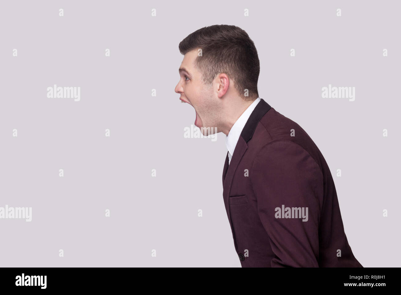 Profile side view portrait aggressive of handsome young man in violet suit and white shirt, standing and screaming with nervous face. indoor studio sh Stock Photo