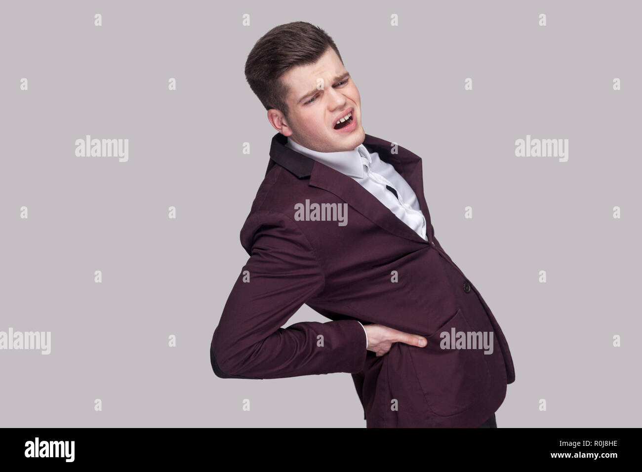 Profile side view portrait of unhappy handsome young man in violet suit and white shirt, standing and holding his painful spine and feeling pain. indo Stock Photo
