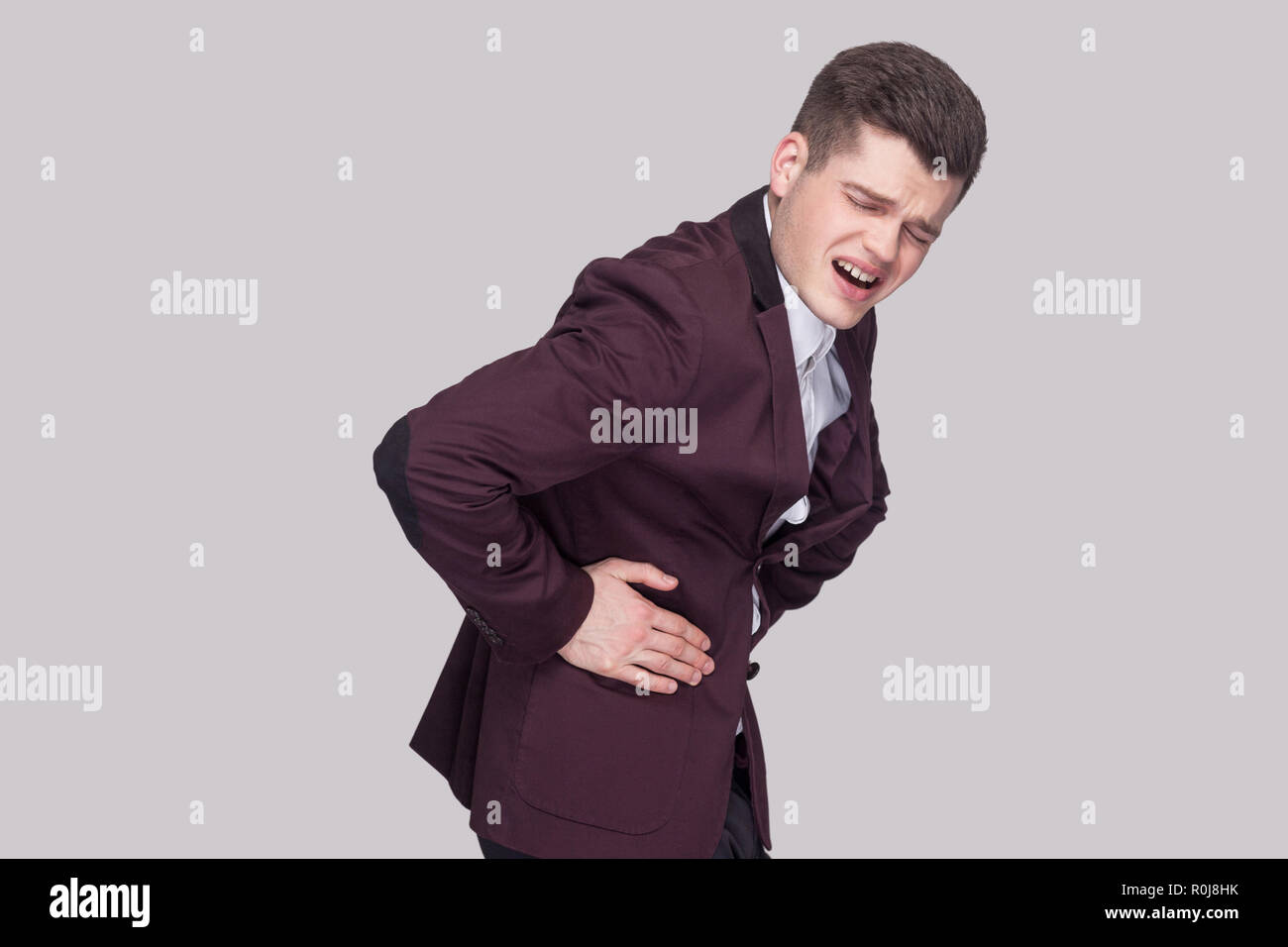 Profile side view portrait of unhappy handsome young man in violet suit and white shirt, standing and holding his painful stomach and feeling pain. in Stock Photo