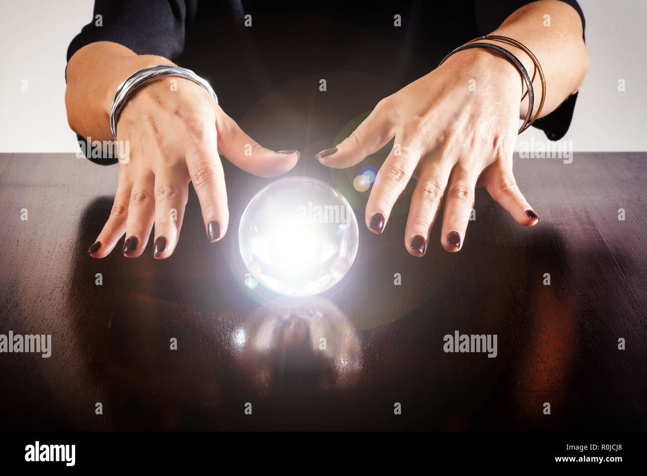 Fortune teller or soothsayer with a glowing crystal ball or sphere reflected on a shiny wooden table in a close up view of her hands Stock Photo
