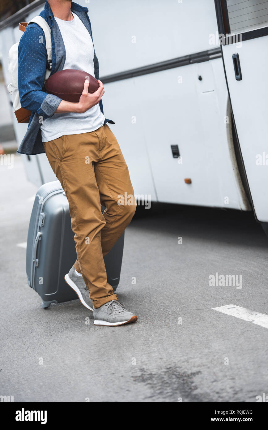 cropped image of man with backpack and rugby ball carrying bag on wheels near travel bus at street Stock Photo