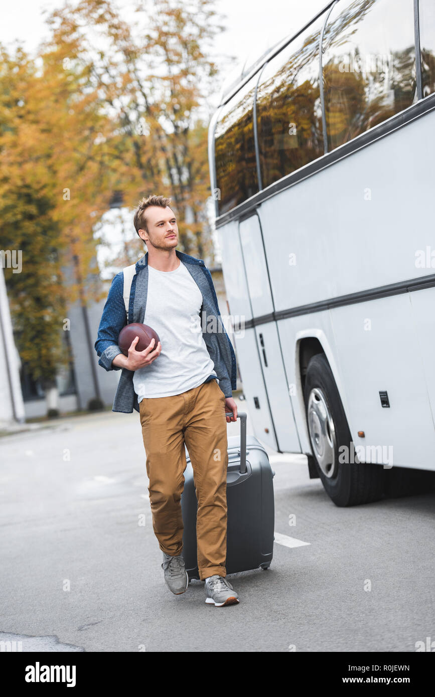 handsome man with backpack and rugby ball carrying bag on wheels near travel bus at street Stock Photo