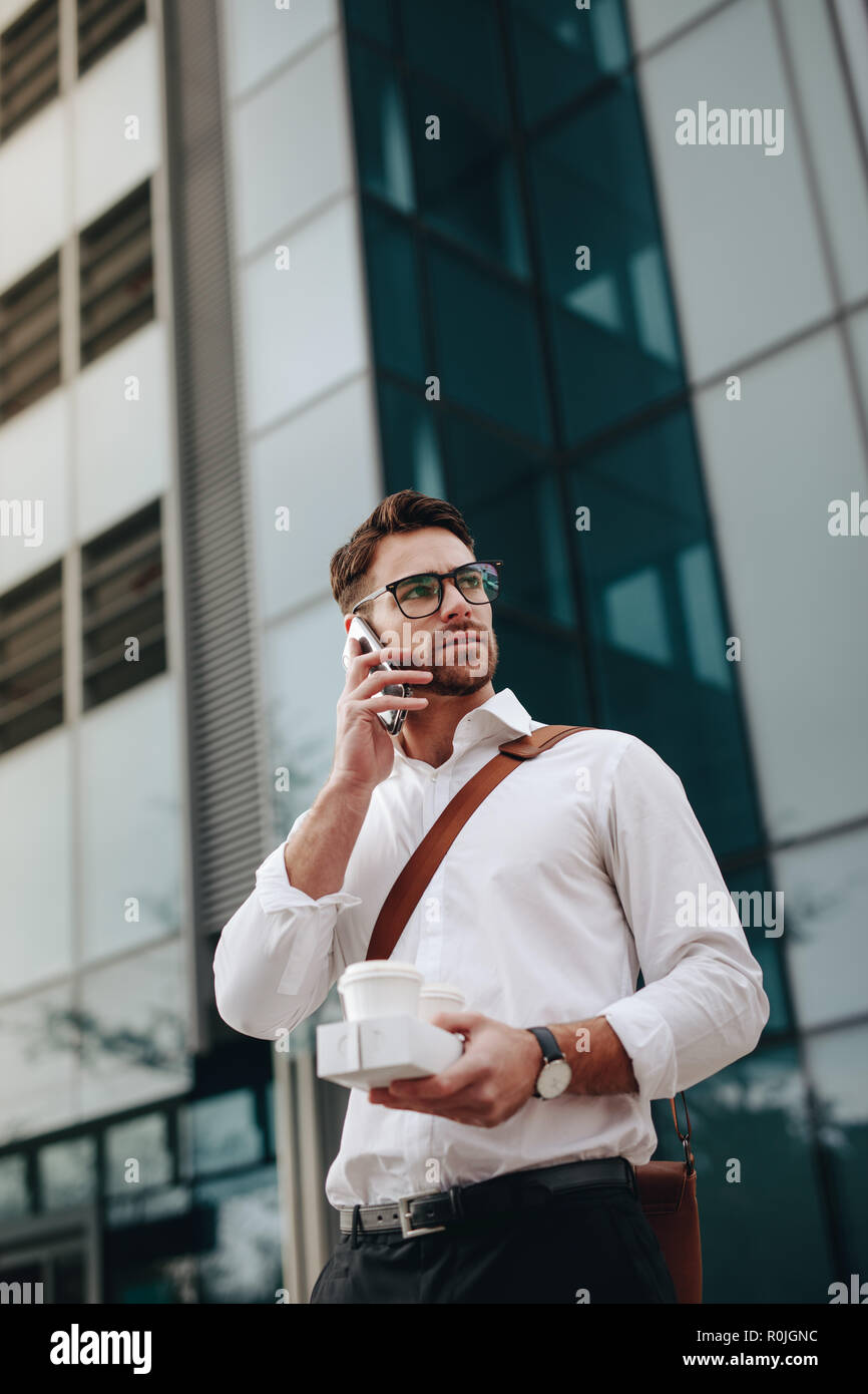 Man carrying two coffee cups in hand talking on mobile phone going to office in the morning. Businessman walking to office talking on mobile phone. Stock Photo