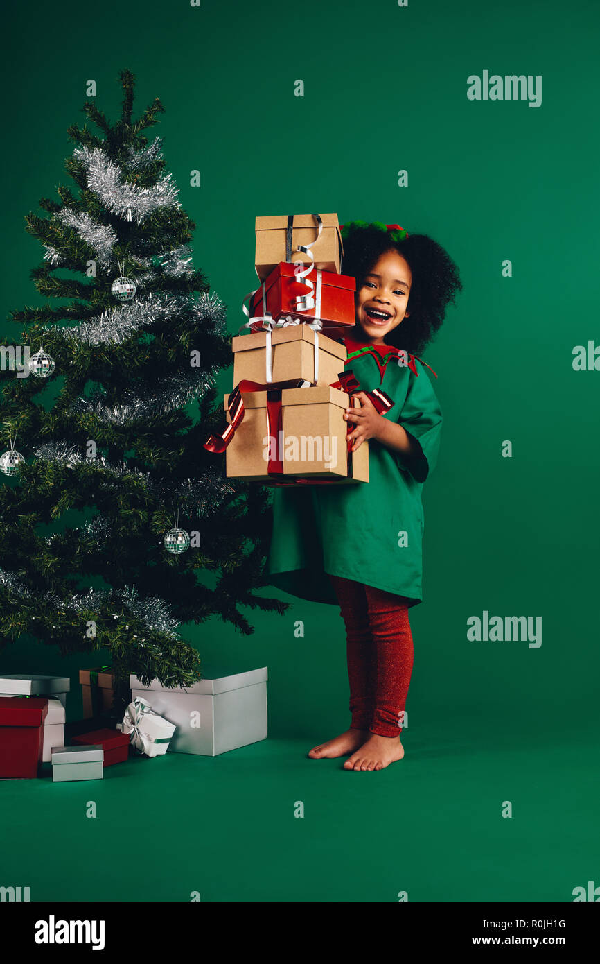 Smiling afro american kid holding her christmas gifts standing beside a christmas tree. Happy kid standing with a pile of christmas gift boxes. Stock Photo